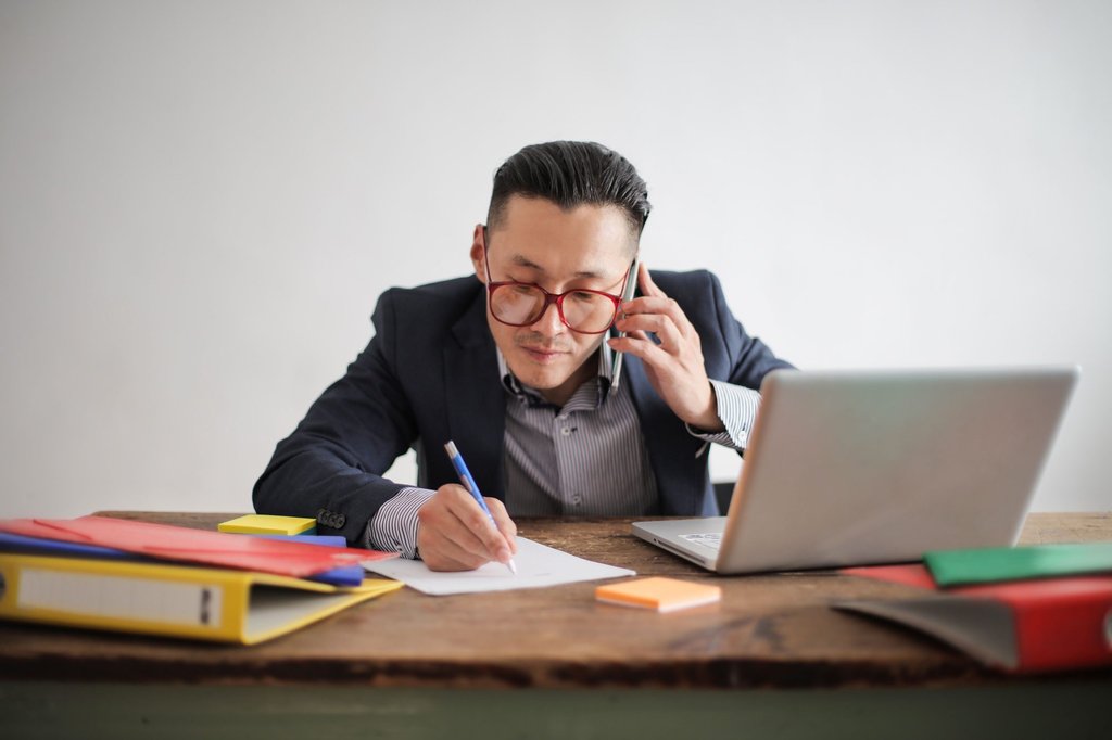 A salesperson taking notes on a call at a desk, with a laptop open beside the notepad.