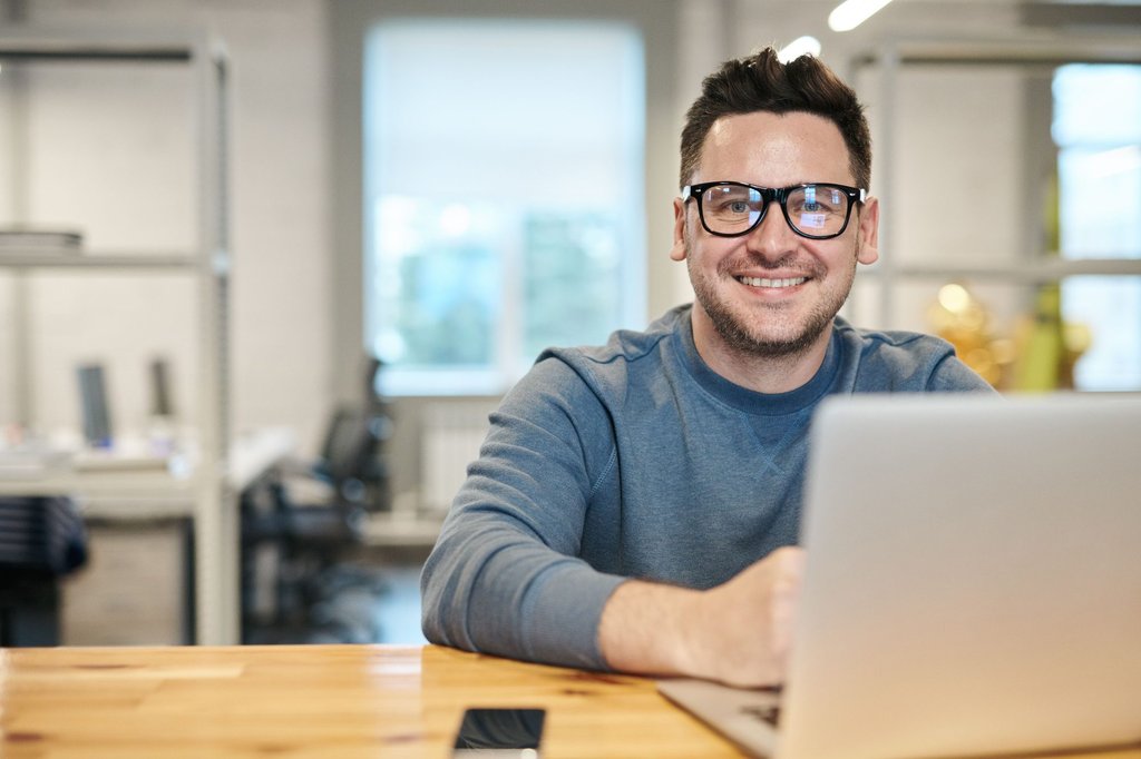 A man in glasses smiling at his laptop in a bright office.