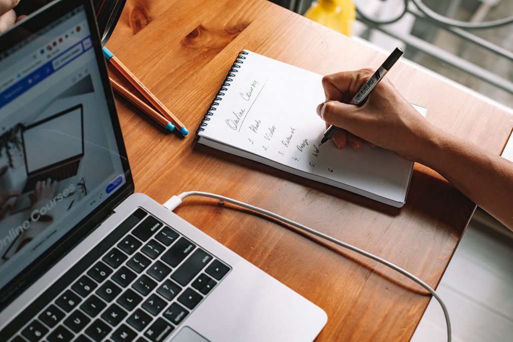 Someone working through an online course on a laptop at home, taking notes as they go.