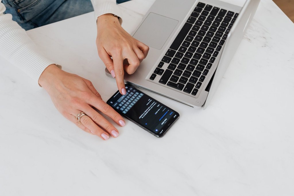Hands using a phone and a laptop together on a white desk, the two devices Zoho Vault keeps credentials in sync across.